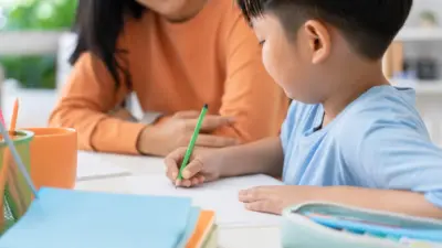 Tangan anak Indonesia menulis di buku dengan orang tua mengamati dengan hangat di meja belajar
